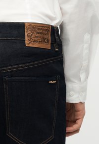 Άτομο που φοράει σκούρο μπλε τζιν με καφέ δερμάτινη ετικέτα της μάρκας και λευκό μακρυμάνικο πουκάμισο, δείχνοντας την περιοχή της πίσω τσέπης.