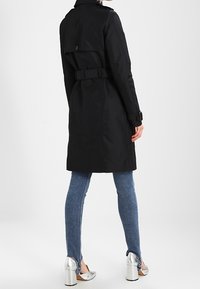 Black trench coat with a belted waist, buttoned back flap, and long sleeves. Worn with denim jeans and silver high-heeled shoes.