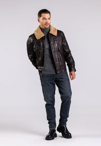 Braune Lederjacke mit einem beigefarbenen Lammfellkragen, vertikalen Nähten und zwei Fronttaschen, kombiniert mit dunkelblauen Jeans und schwarzen Stiefeln.