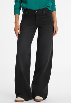 Femme portant un jean noir à jambes larges, un haut vert à manches longues et des baskets blanches, debout les mains détendues le long du corps.