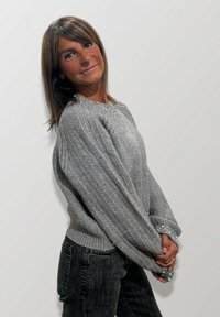 Donna con capelli castani che indossa un maglione grigio a maglia con dettagli di perline e jeans neri, mentre posa di lato contro uno sfondo chiaro e semplice.