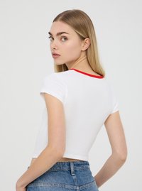 T-shirt bianco accorciato con maniche corte e scollatura rossa. Il materiale sembra morbido e leggero. Il modello indossa pantaloni shorts in denim a vita alta.