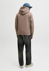 Φούτερ με κουκούλα σε ανοιχτό καφέ χρώμα, από μείγμα βαμβακιού με ριμπ μανσέτες· συνδυασμένο με φαρδιά, ξεθωριασμένα μαύρα τζιν και μπεζ αθλητικά παπούτσια. Ανοιχτά μπλε καπέλο φορεμένο ανάποδα.