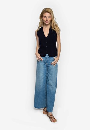 Blondine staand met handen in de zakken, gekleed in een mouwloze zwarte button-down vest, wijde blauwe jeans en bruine sandalen met bandjes.