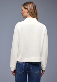 Frau von hinten gesehen, die ein strukturiertes weißes Langarmshirt und blaue Jeans vor einem einfarbigen grauen Hintergrund trägt.