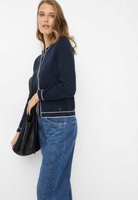 Marineblå cardigan med hvide pipingdetaljer, lange ærmer og knapdetaljer. Kombineret med lyseblå jeans og en sort håndtaske, sat mod en hvid baggrund.
