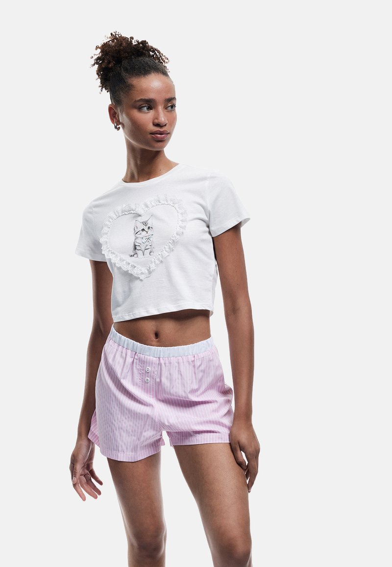 Hvid cropped t-shirt med en kattegrafik og et yndigt hjerte-design med flæser; sat sammen med lyserøde stribede shorts med elastisk talje.