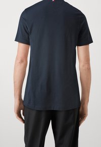 Tmavomodré bavlněné tričko s krátkým rukávem, kulatým výstřihem a malým červeným a bílým pruhem na zadní straně límce.