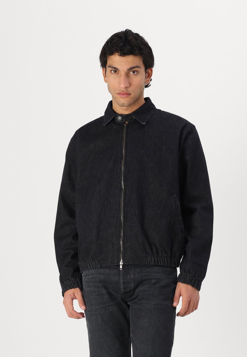 Zwart denim jack met een ritssluiting aan de voorkant, drukknopenkraag, elastische onderkant en zijzakken, gedragen met bijpassende zwarte jeans.
