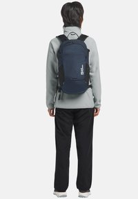Zaino blu navy con una texture liscia, dotato di pannelli laterali in rete e spallacci regolabili, indossato sopra un pile grigio chiaro e pantaloni neri.