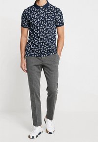 Homme portant un polo floral bleu marine, un pantalon slim gris et des baskets blanches, se tenant avec une main dans sa poche.