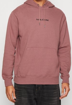 Persona che indossa una felpa Volcom rosa polvere con tasca frontale e coulisse, abbinata a jeans scuri.