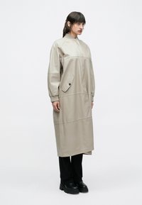 Žena v dlhom béžovom kabáte s zapínateľnými vreckami, čiernych nohaviciach a čiernych robustných topánkach, stojaca pred jednoduchým bielym pozadím.