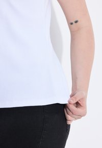 Top a canottiera bianca a costine con una forma aderente, con le cuciture laterali in vista. Una mano afferra l'orlo, in contrasto con i pantaloni neri.