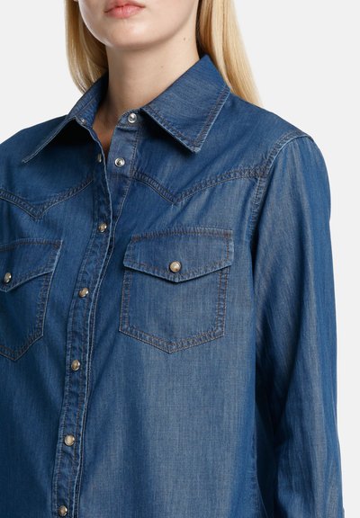 Denim srajca v temno modri barvi, z razporkom na ovratniku, stiskalnimi gumbi in dvema žepoma na prsih z pokrovoma. Gladka tekstura z diskretnimi šivi.