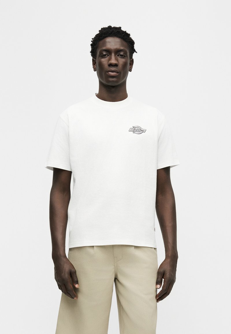 Homme portant un t-shirt blanc à manches courtes avec un petit logo sur la poitrine et un pantalon beige, debout devant un fond uni gris clair.