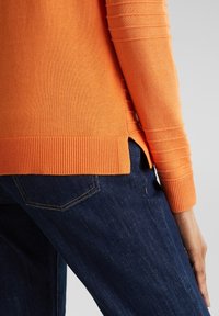 Oranger Strickpullover mit gerippter Textur, ausgestattet mit langen Ärmeln und einem gespaltenen Saum, kombiniert mit dunkelblauen Jeans.