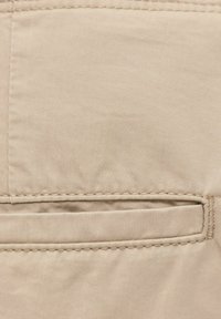 Gros plan sur un tissu beige montrant une poche passepoil horizontale avec des coutures détaillées sur un vêtement.