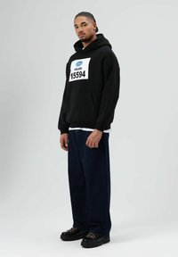 Zwarte hoodie met een wit label waarop "POLIZEI 15594" staat, gecombineerd met donkerblauwe oversized jeans en zwarte plateau schoenen met rode veters.