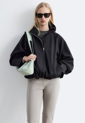 Femme portant une veste noire zippée oversize, pantalon beige clair, lunettes de soleil noires, boucles d’oreilles argentées en forme d’anneau, bagues, et tenant un sac à main vert pâle.