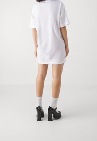 Osoba ubrana w białą, oversize'ową sukienkę typu T-shirt, białe skarpetki do kostek oraz czarne, na wysokim obcasie buty typu Mary Jane, stoi plecami do białego tła.