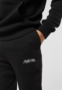 Detail osoby v černých teplácích s rukou v kapse a bílým nápisem "CLSR LDN Design Atelier" na stehně.