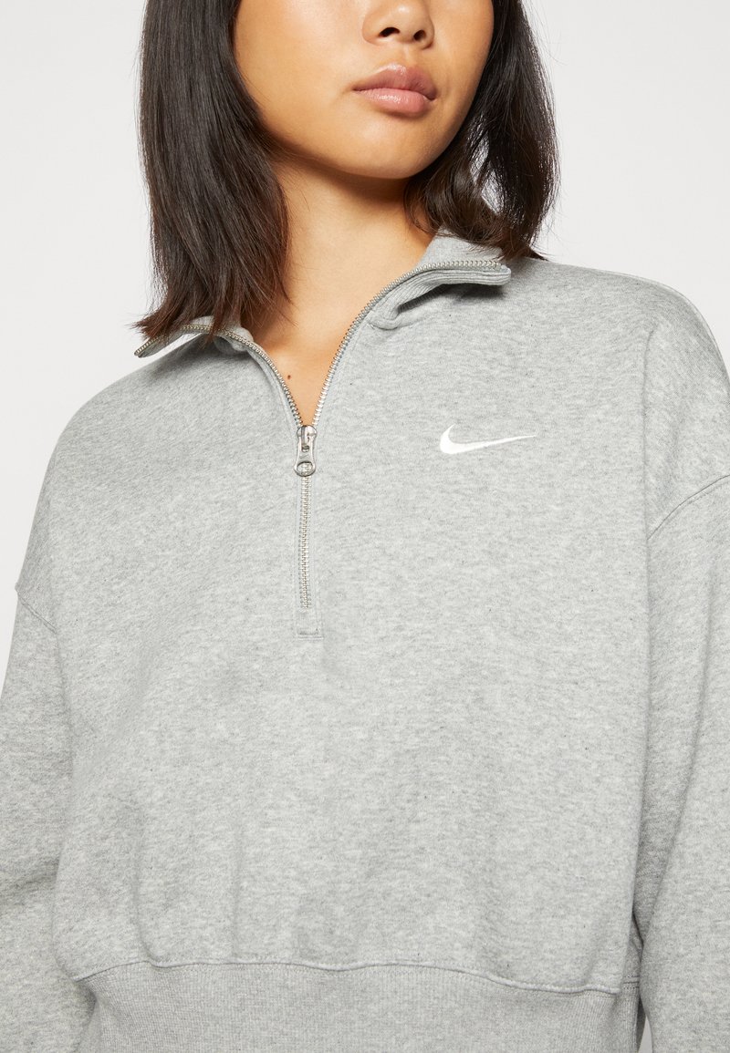 Sudadera gris con cremallera y cuello alzado, con un pequeño logo blanco de Nike. La tela es suave, con un corte holgado y ribetes en el bajo.