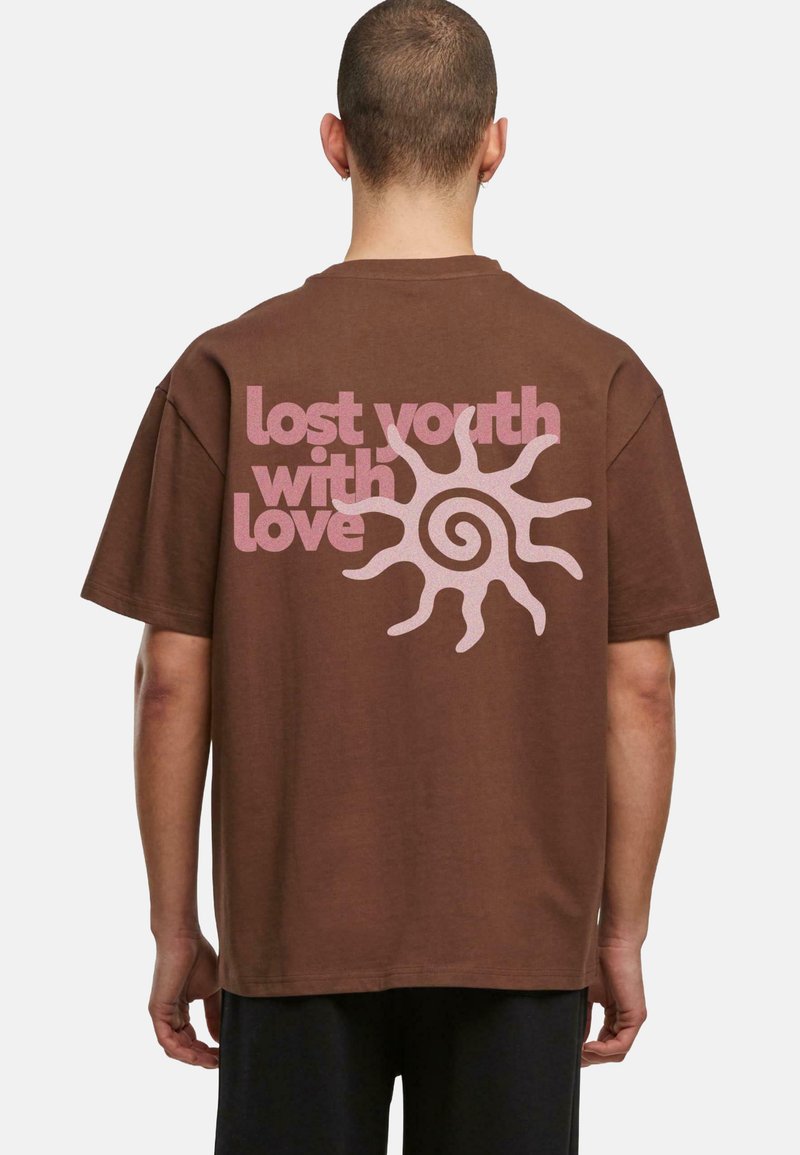 Hnědé oversized tričko s růžovým textem a designem slunce na zádech, vyrobené z bavlny s hladkou texturou. Krátké rukávy.