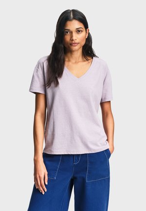 Femme aux longs cheveux bruns portant un t-shirt violet clair à col en V et manches courtes, et un pantalon bleu, debout avec une main dans la poche.