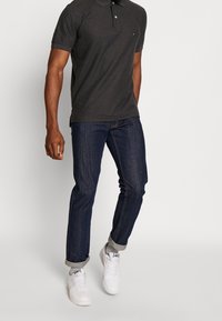 Polo shirt gris foncé à manches courtes et col, associé à un jean bleu foncé à revers et des baskets blanches. Design simple et décontracté.