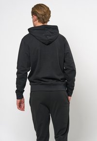 Black hoodie with a kangaroo pocket, ribbed cuffs and hem, and a drawstring hood. Soft, textured fabric, model shown from the back.
