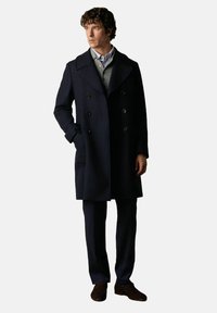 Námornická vlněná peacoat s dvouřadým designem, širokými klopami a bočními kapsami, spárovaná se svetrem světle šedé barvy a tmavými kalhotami.