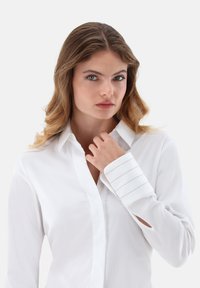 Camicia bianca con collo a punta e texture liscia, caratterizzata da dettagli a strisce sui polsini. Maniche lunghe con ritagli decorativi.