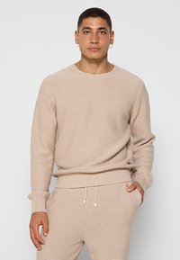 Junger Mann in beigem, strukturiertem Strickpullover und passender Jogginghose, steht mit einer Hand in der Tasche vor einem schlichten Hintergrund.