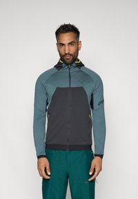 Kapucnis, sötétszürke és türkiz anyagú, kontrasztos sárga részletekkel ellátott zipzáras dzseki, oldalsó zsebekkel és ergonómikus kialakítással.