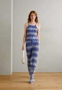 Jumpsuit à motifs zigzag bleu et blanc avec un design col halter. Le mannequin tient un sac à main rose clair. Tissu doux, silhouette ajustée.
