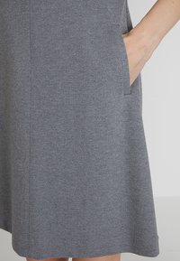 Main reposant dans la poche droite d'une robe en tricot gris longueur genou avec des détails de couture visibles.