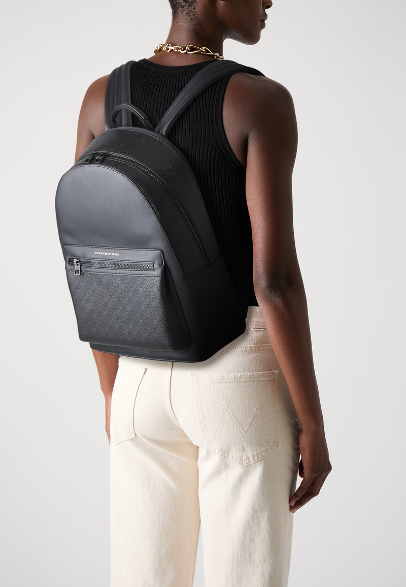 Black leather backpack with textured front, curved shape, and a zippered pocket. Adjustable straps and smooth finish. Neutral-toned pants visible.