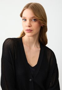 Cardigan nero a maglia aperta con maniche ampie, scollatura a V e un singolo bottone. Indossato sopra un top nero, caratterizzato da un tessuto testurizzato.