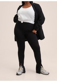 Leggings noirs taille haute avec fentes sur les côtés, associés à un débardeur blanc noué et un long blazer noir. Des baskets grises et blanches complètent le look.