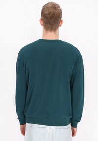 Felpa teal con maniche lunghe, scollatura rotonda e polsini a coste. Tessuto morbido con una trama liscia, dettagli minimi e vestibilità rilassata.