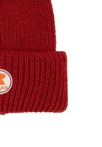 Red knitted beanie with a folded cuff and a circular logo patch featuring an orange design on a white background. Textured, warm material.