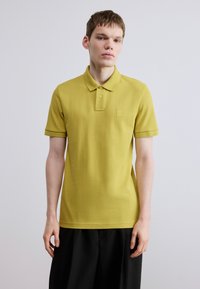 Polo jaune moutarde, manches courtes, col classique, tissu texturé, avec une petite étiquette de marque sur le côté gauche de la poitrine.