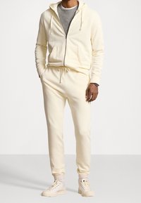 Ljus krämfärgad huvtröja med dragkedja, kombinerad med matchande joggers som har dragsko i midjan och ribbade muddarna. Bärs med tanfärgade hög- topp sneakers.