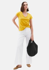 T-shirt jaune à col en V, pantalons blancs à jambes larges, tenant un sac tissé noir. Présente un matériau texturé et des sandales ouvertes.