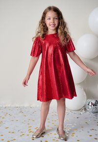 Rød palietkjole med korte puffærmer og en vid nederdel. Kombineret med guld sko. Baggrunden prydes af balloner og konfetti.