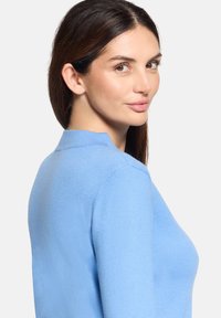 Kobieta o długich ciemnych włosach, ubrana w jasnoniebieski sweter, spoglądająca przez ramię na jednolitym białym tle.