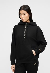 Νεαρή γυναίκα που φοράει μαύρο φούτερ με κουκούλα EA7 Emporio Armani και αντίστοιχο παντελόνι με λεπτομέρειες λογότυπου, στέκεται με το χέρι στην τσέπη, ουδέτερο φόντο.