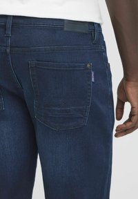 Tmavomodré džíny z denimu s hladkou textúrou, s zadnou kapsou so šitím v tvare hory a malým štítkom.