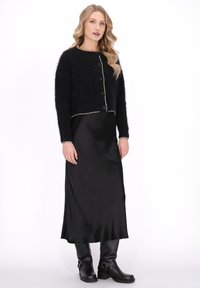 Vrouw met lang blond haar, gekleed in een zwarte fluwelen knopen cardigans over een zwarte satijnen jurk en zwarte kniehoge laarzen, staand tegen een witte achtergrond.